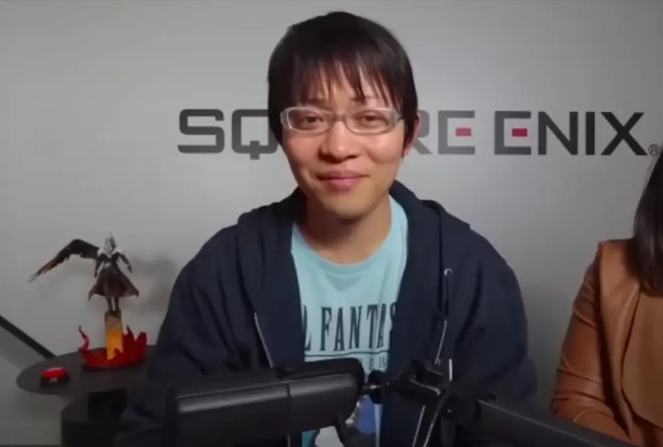 Naoki Hamaguchi w okularach i niebieskim t-shircie, siedzący na tle ściany z logo Square Enix.