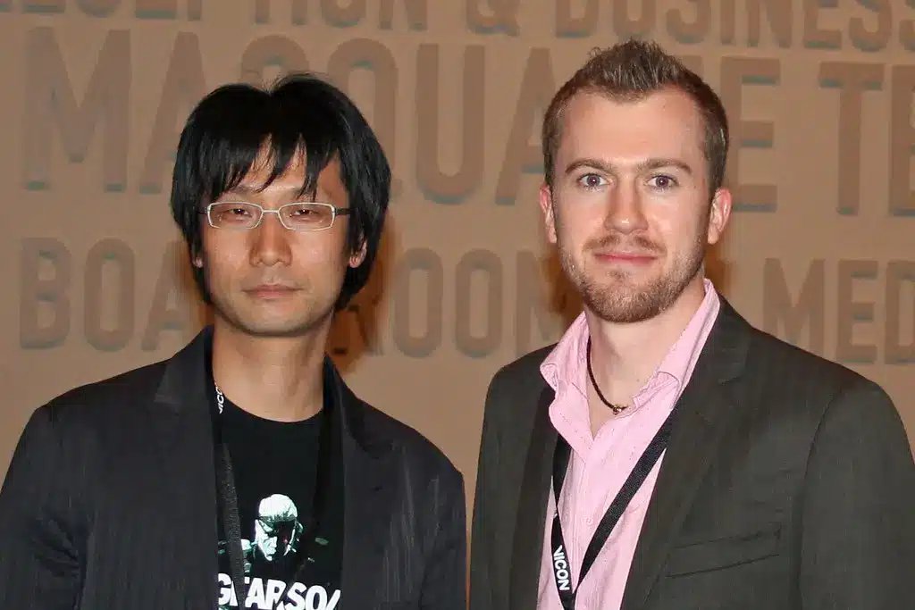 Hideo Kojima w okularach i czarnej marynarce, stojący obok innego mężczyzny.